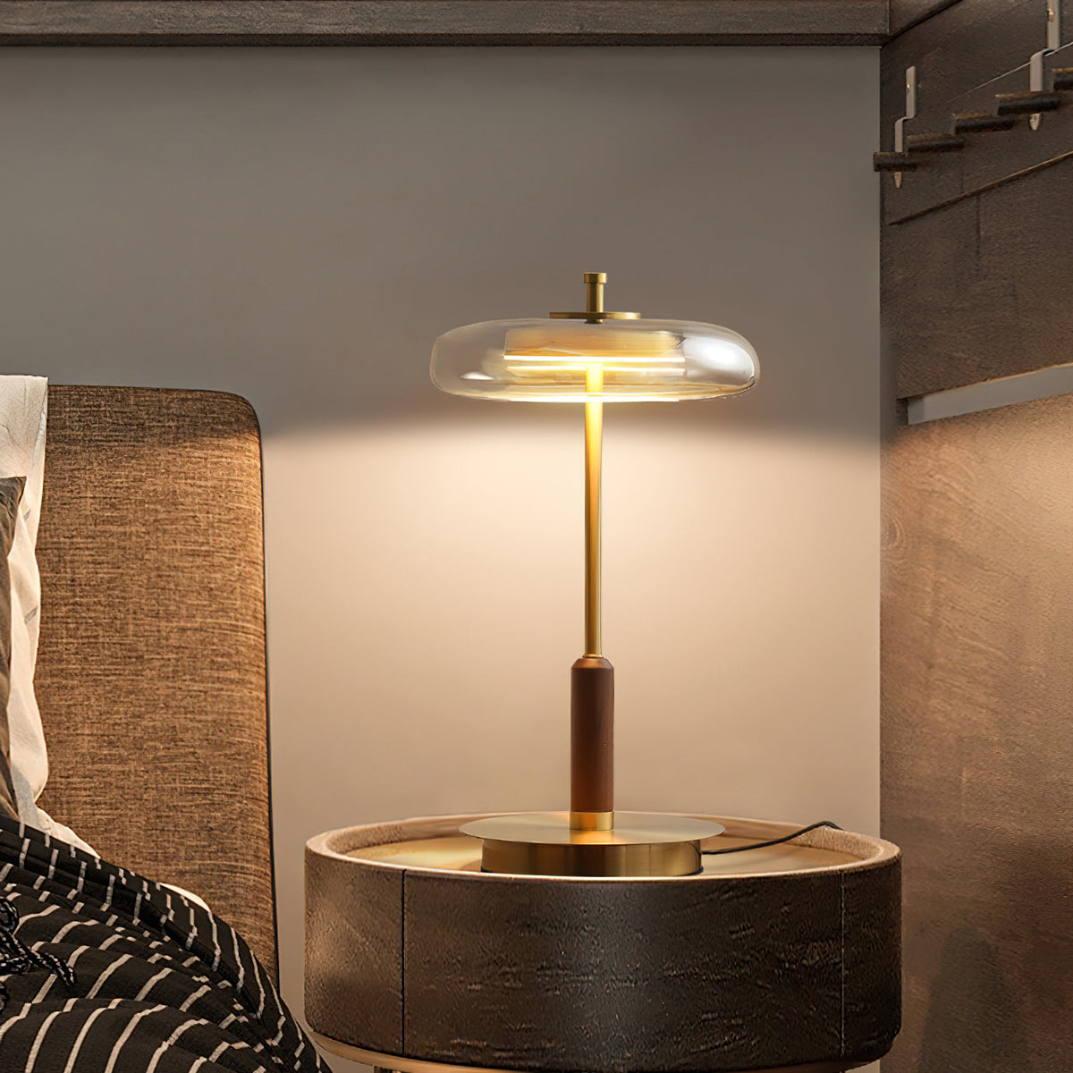 Clérachic table lamp glowing on a round bedside table, the brass base and walnut stem set against a woven headboard.