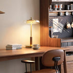 Walnut desk corner with a Clérachic table lamp switched on, books and a cup at its base and a pegboard shelf behind.