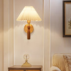 Lit Clérachic wall light with a pleated cream shade on a pale panelled wall, above a small chest topped with a glass ornament.