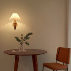 A Clérachic pleated wall light glowing on a warm beige wall above a round wood table with a vase of green stems and a rust-coloured chair.