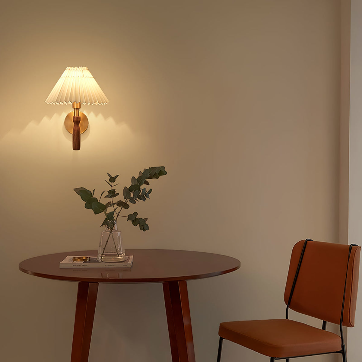 A Clérachic pleated wall light glowing on a warm beige wall above a round wood table with a vase of green stems and a rust-coloured chair.