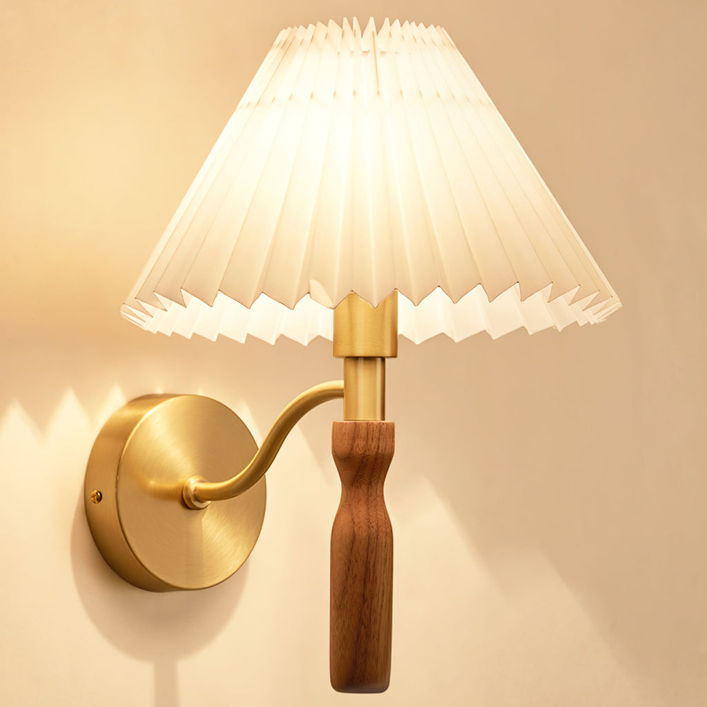 Close view of this Clérachic wall light lit, the pleated shade glowing above a walnut grip and an S-curved brass arm.