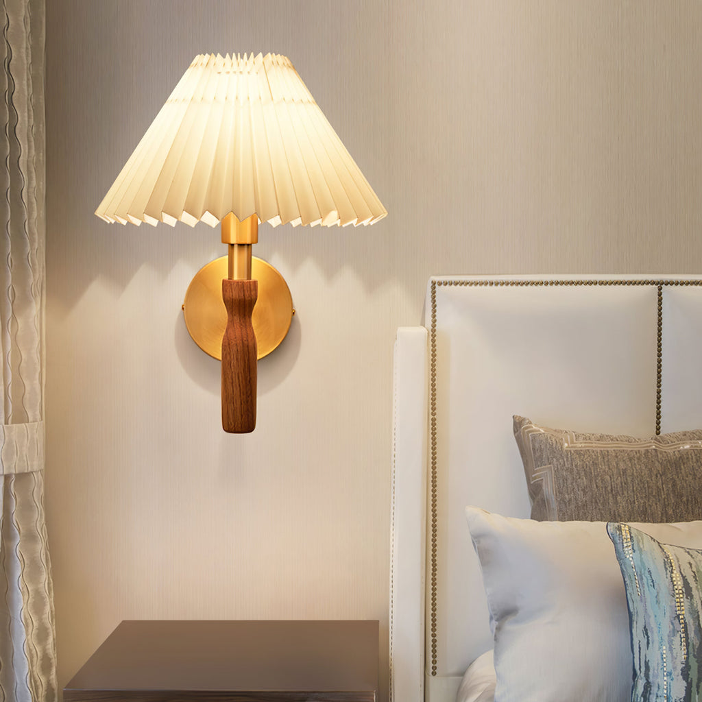 A Clérachic wall light lit beside an upholstered bed head, its pleated shade and brass plate against a warm beige wall.