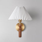 Front view of a pleated white Clérachic wall light unlit on a pale grey wall, with a walnut stem and round brass back plate.