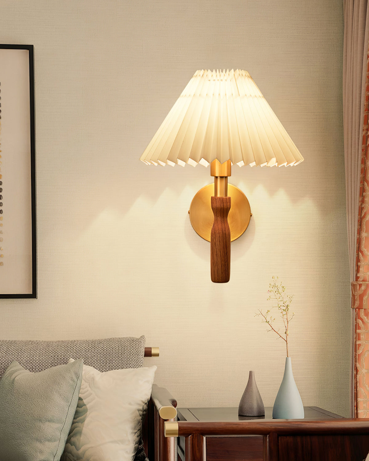 Lit Clérachic wall light with a cream pleated shade on a warm wall above a bed, with two slim vases on the bedside table.