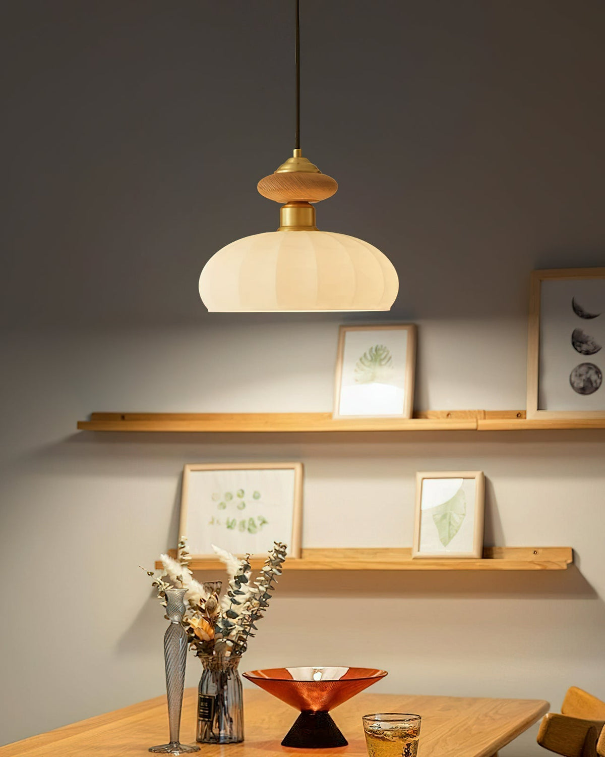 This Clérachic pendant casts a pool of light on a grey wall with two shelves of small framed prints and a vase of dried stems.