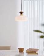 A Clérachic pendant with a ribbed glass shade hangs beside sheer white curtains, above a table with a clay pot and a stack of books.