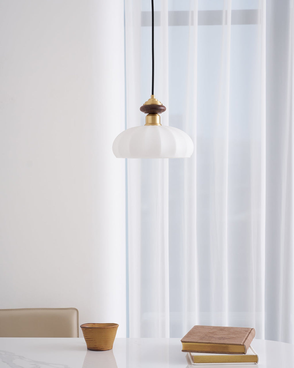 Clérachic pendant with a walnut and brass top and a ribbed white shade, in front of floor-length sheer curtains above a marble table.