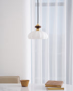 Clérachic pendant with a walnut and brass top and a ribbed white shade, in front of floor-length sheer curtains above a marble table.