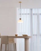 A smaller view of a Clérachic pendant lit above a pale pedestal table with a leather-look chair and window curtains behind.