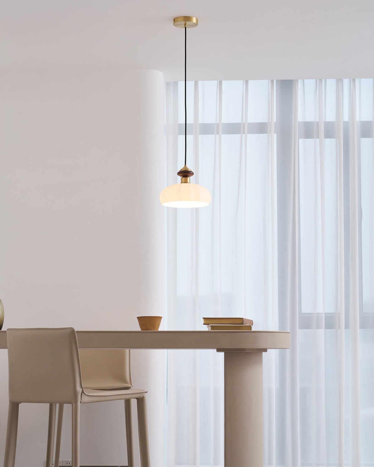 A smaller view of a Clérachic pendant lit above a pale pedestal table with a leather-look chair and window curtains behind.