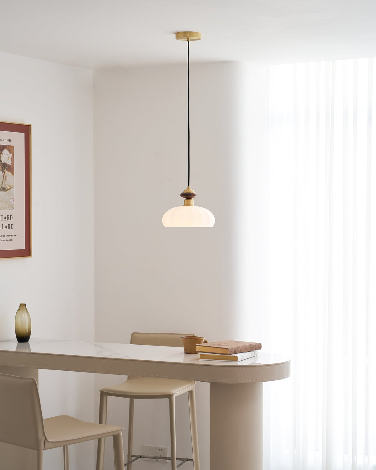 Wide view of a Clérachic pendant with a glowing ribbed shade hung over a pale table and bar stools beside a framed print.