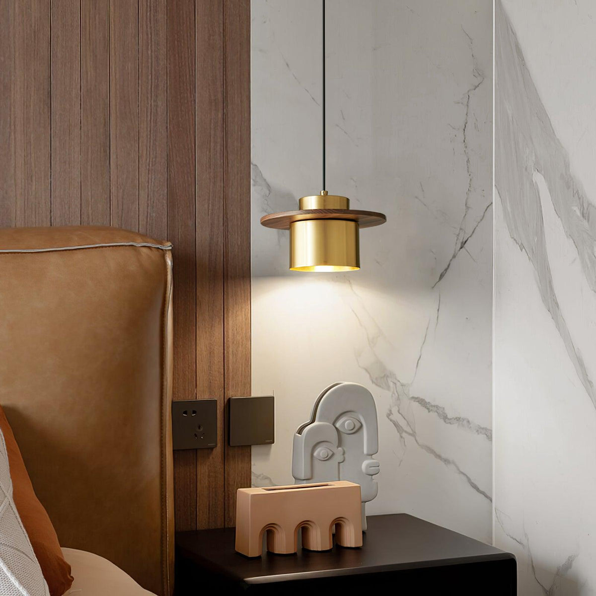 A brass-and-walnut Clérachic pendant glows beside a bed with a tan leather headboard, white marble and a pink ceramic ornament.
