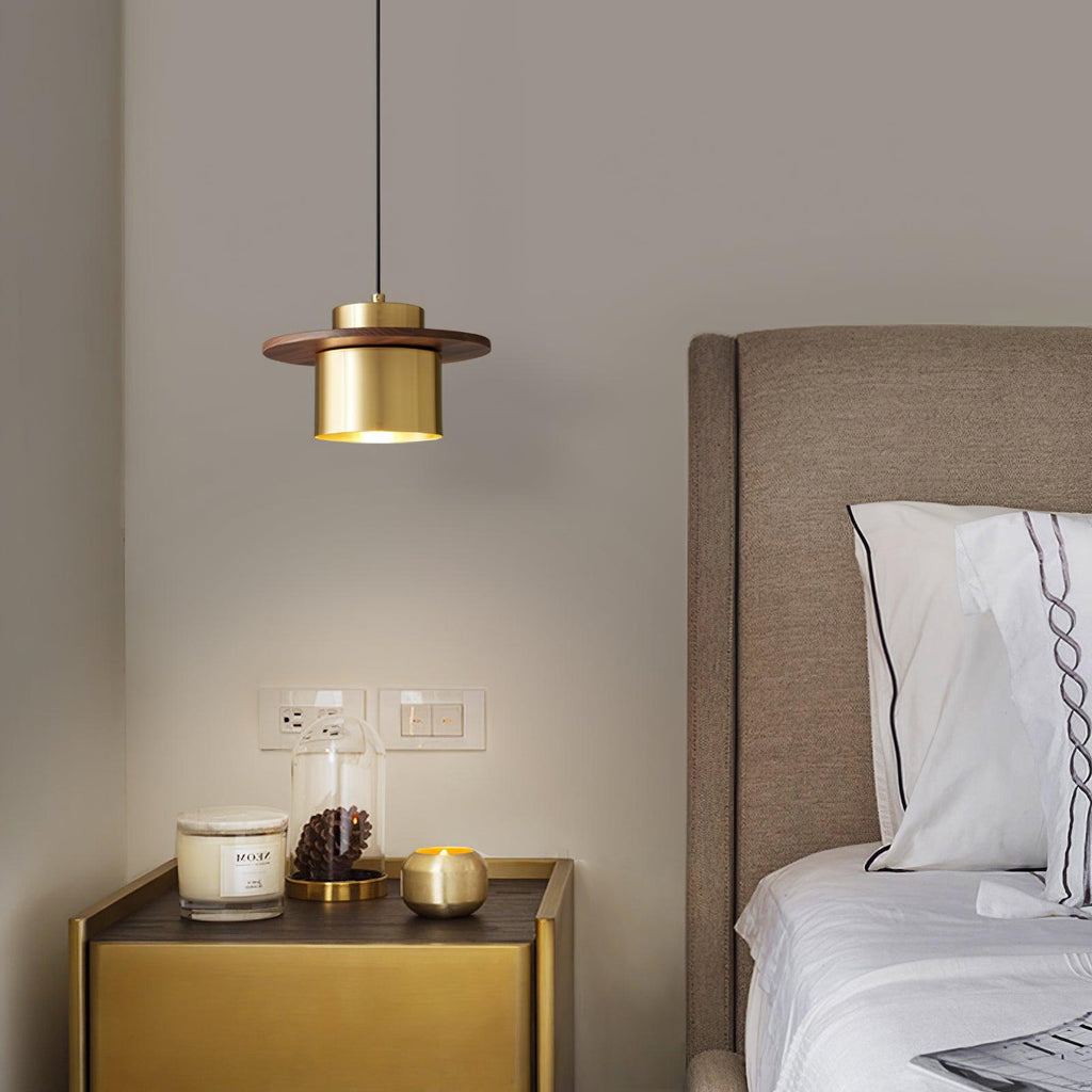 Small Clérachic pendant with a brass cylinder and walnut brim hangs above a gold bedside table with a candle and a bed beyond.