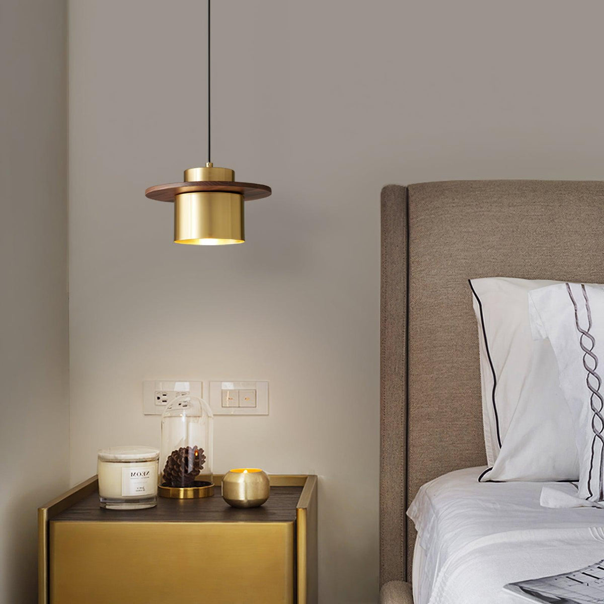Small Clérachic pendant with a brass cylinder and walnut brim hangs above a gold bedside table with a candle and a bed beyond.