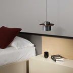 Black-and-walnut Clérachic pendant hangs above a cream bedside shelf with a black cup and books, beside a bed with a red cushion.