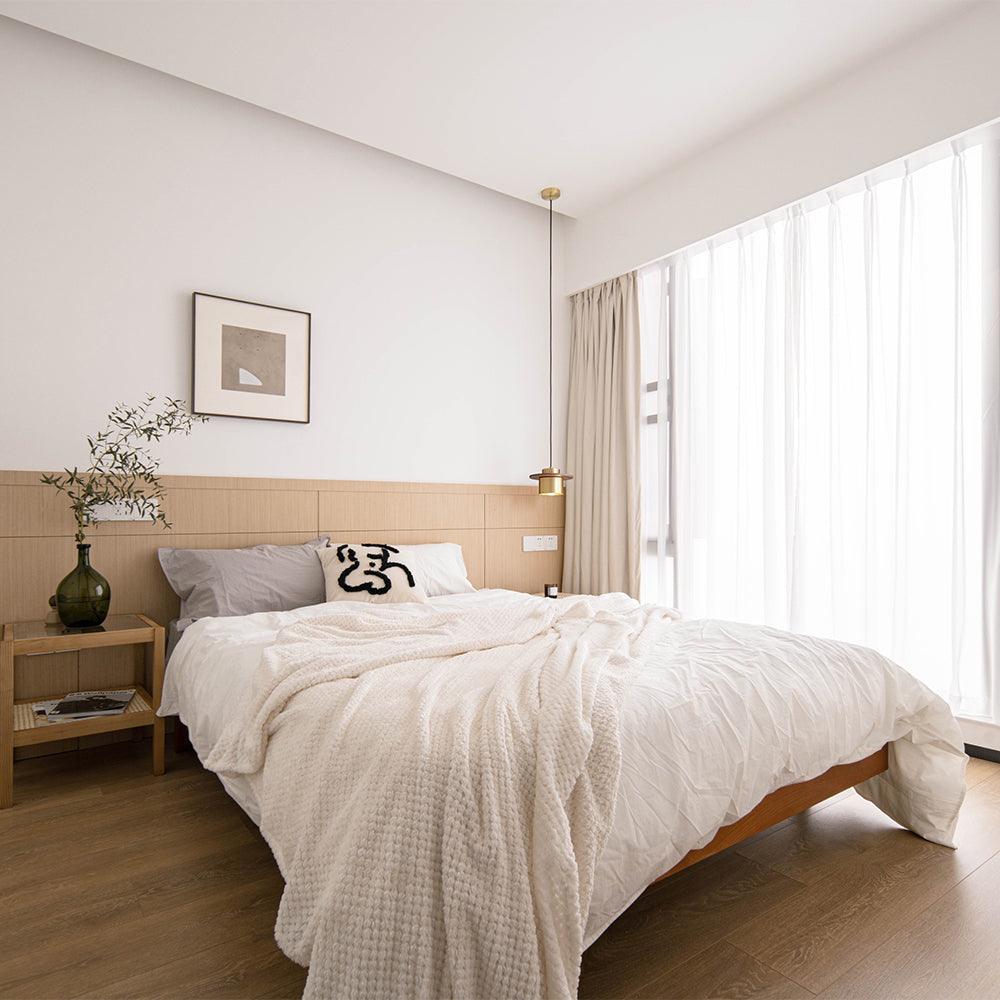 A small brass Clérachic pendant hangs by a bed with white linen, a wooden headboard panel and floor-length sheer curtains.