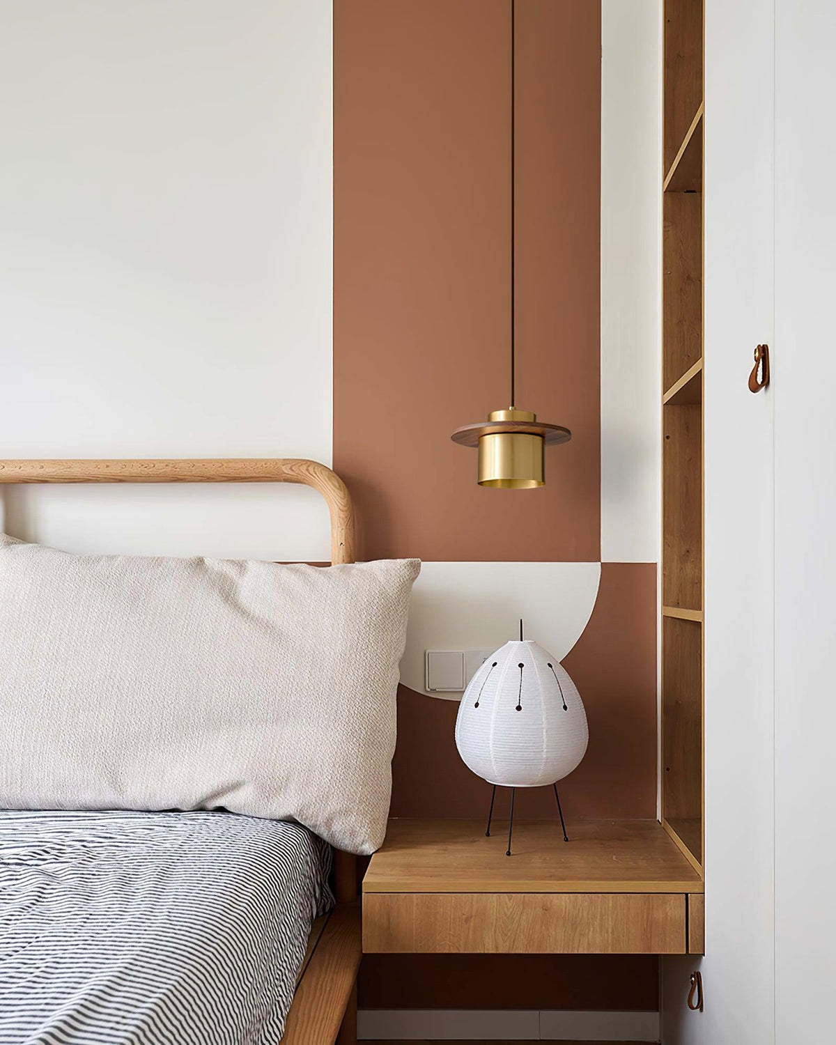 A Clérachic pendant with a brass shade hangs by a bed with a rattan headboard, an orange wall panel and a paper lamp.