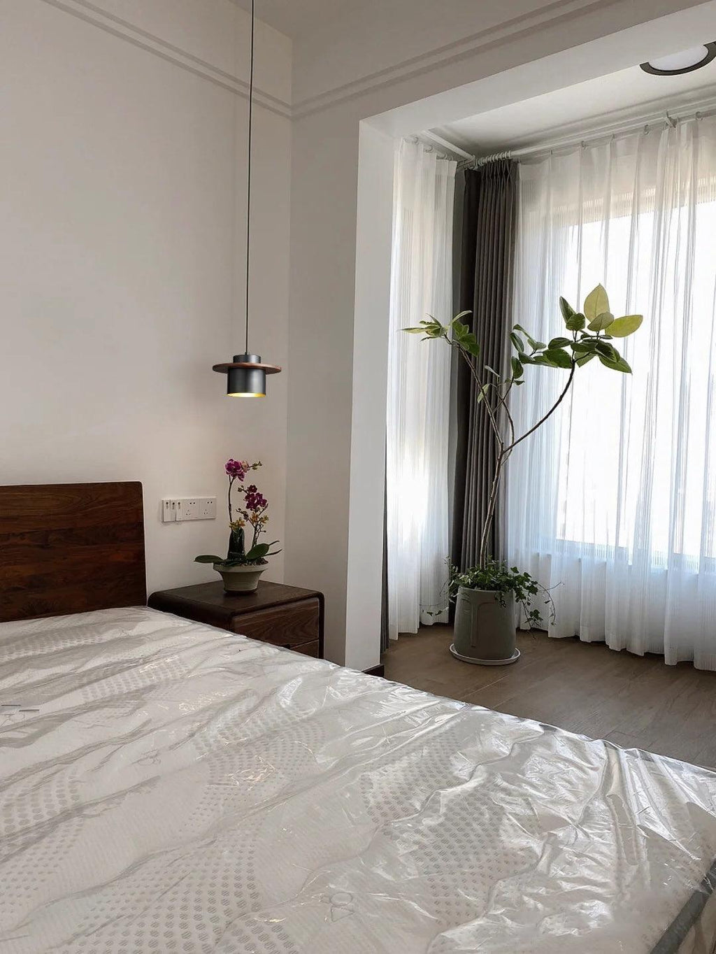 A small black-and-walnut Clérachic pendant hangs near a bed with a dark wood headboard, with tall sheer curtains and a plant.