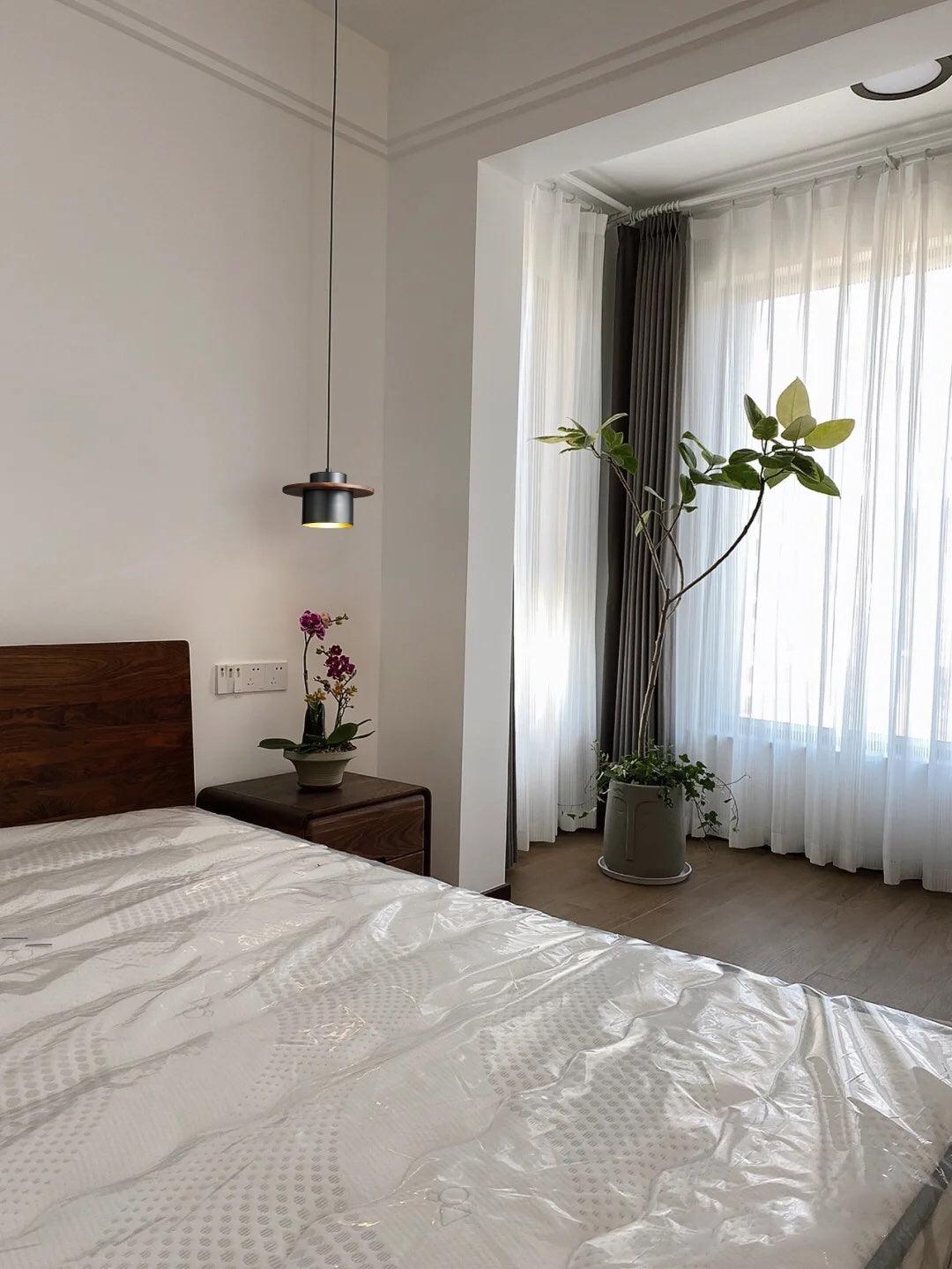 A small black-and-walnut Clérachic pendant hangs near a bed with a dark wood headboard, with tall sheer curtains and a plant.
