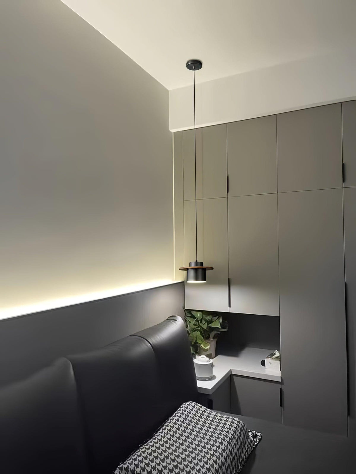 A Clérachic pendant with a dark shade hangs above a bed in a grey room with wall cabinets and a strip of warm light.