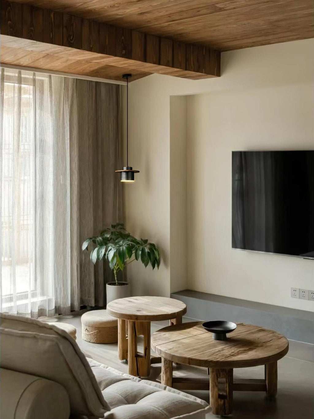 A tiny dark Clérachic pendant hangs in a living room with a wood-beamed ceiling, a round wooden table and a wall-mounted TV.