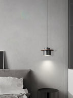 Black-and-walnut Clérachic pendant glows beside a grey bed in a pale room, with a ribbed black stool below.