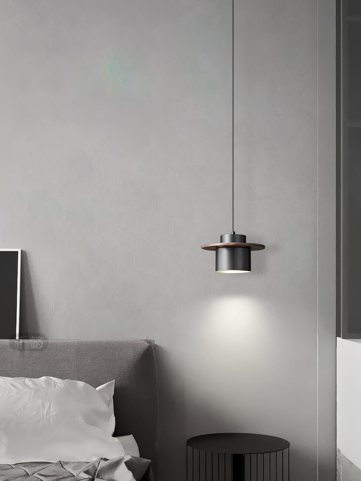 Black-and-walnut Clérachic pendant glows beside a grey bed in a pale room, with a ribbed black stool below.