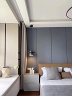A black-and-walnut Clérachic pendant hangs beside a bed with a tan leather headboard and a grey wall panel behind.