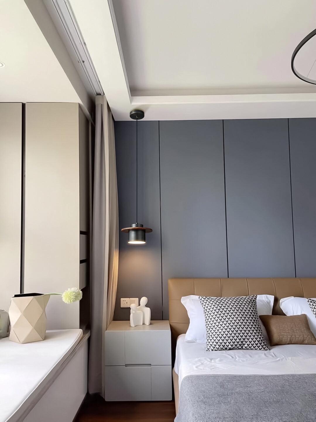 A black-and-walnut Clérachic pendant hangs beside a bed with a tan leather headboard and a grey wall panel behind.