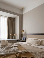 A single Clérachic pendant with a brass cylinder hangs beside a bed with a rattan headboard and beige curtains.