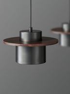 Black cylinder shade under a round walnut brim on this Clérachic pendant, with a second pendant softly blurred behind.