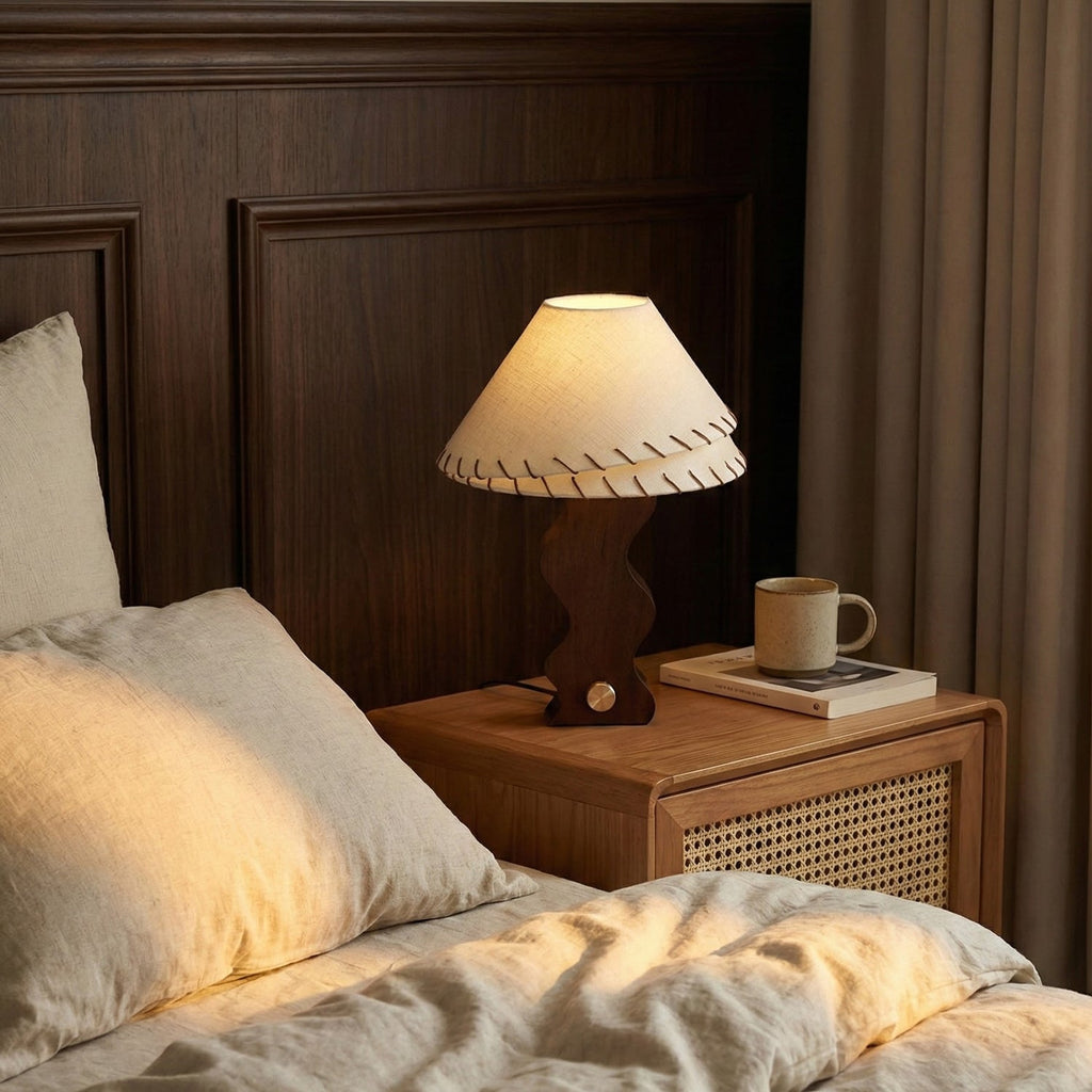 Lit Clérachic table lamp on a rattan bedside table, its linen shade glowing beside a mug, a book and rumpled linen bedding.