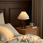 Lit Clérachic table lamp on a rattan bedside table, its linen shade glowing beside a mug, a book and rumpled linen bedding.