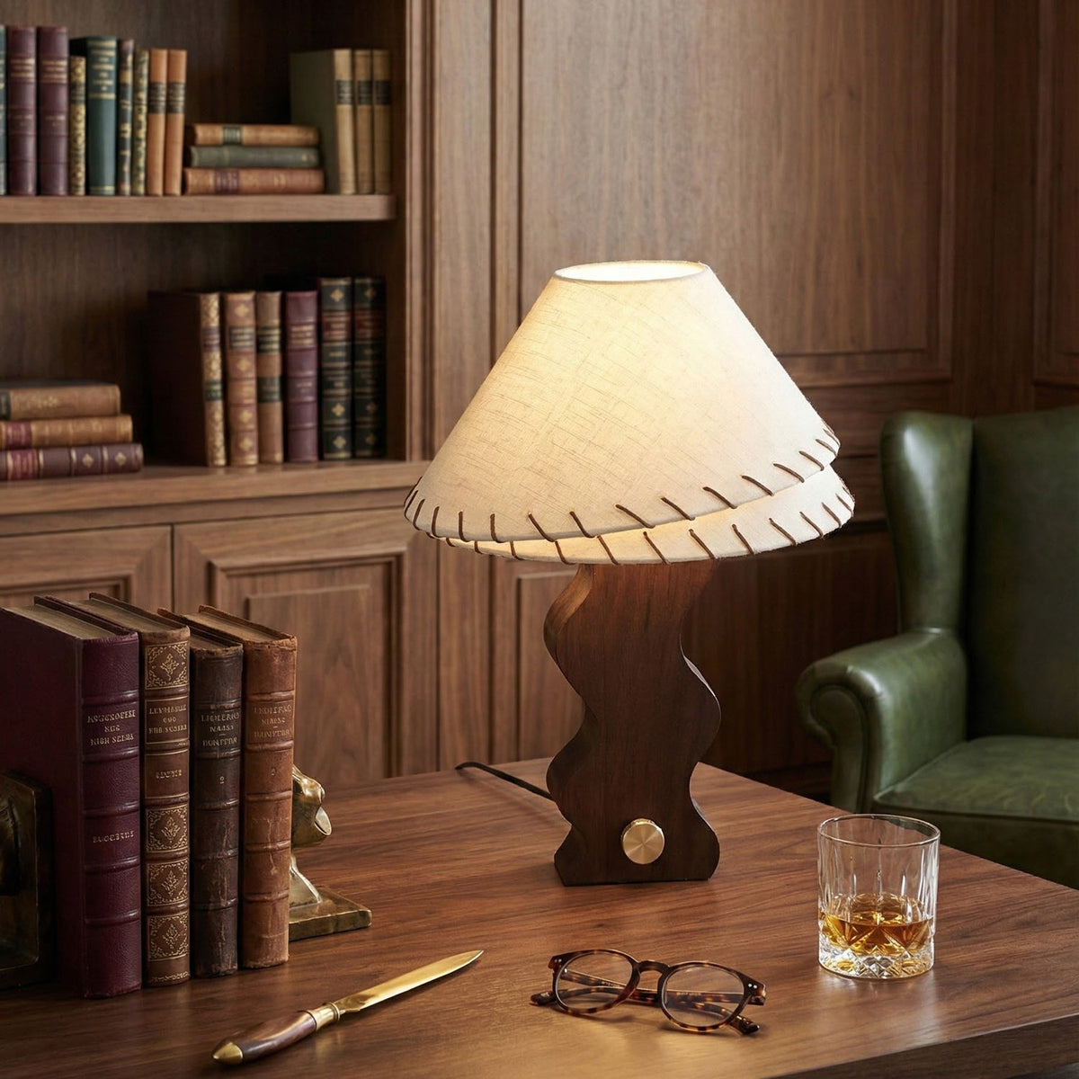 Clérachic walnut table lamp on a dark wooden desk, lit, with leather-bound books, a pair of glasses and a glass of whisky.