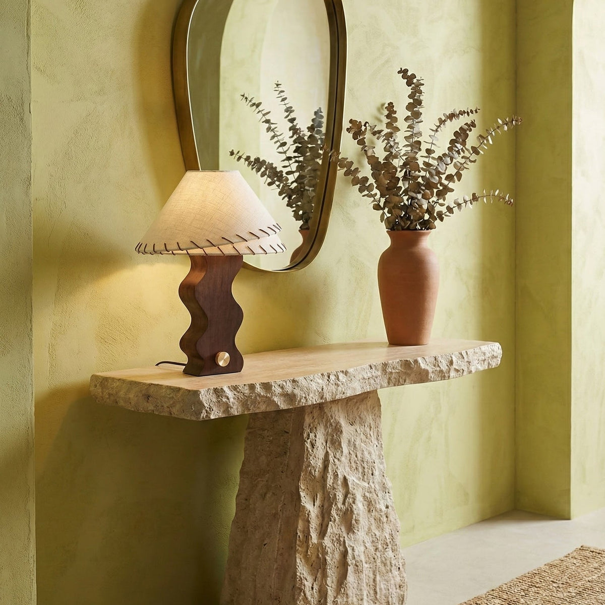 Small Clérachic table lamp on a rough stone console under an arched mirror, next to a vase of dried eucalyptus.
