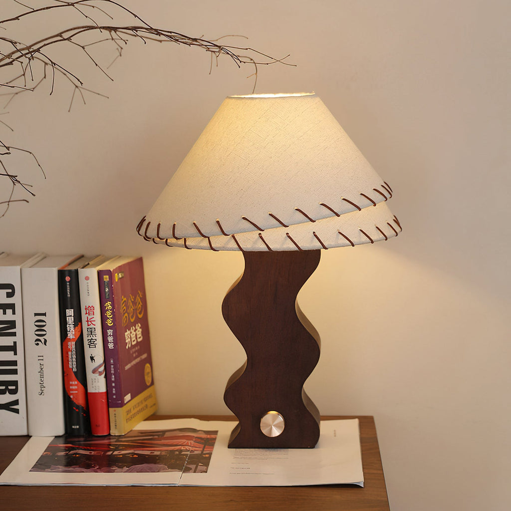 Lit walnut table lamp from Clérachic on a wooden shelf, with a stack of books and a magazine beside the wavy base.