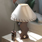 Close view of a Clérachic table lamp's laced linen shade and wavy walnut base on a travertine block with a reed diffuser.