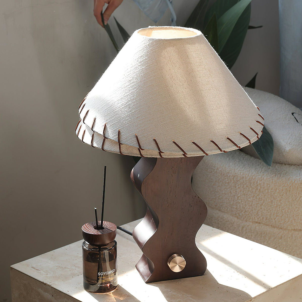 Close view of a Clérachic table lamp's laced linen shade and wavy walnut base on a travertine block with a reed diffuser.