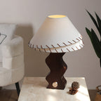 White linen shade with leather stitching on a Clérachic walnut lamp, set on stone beside a pine cone and a cream armchair.
