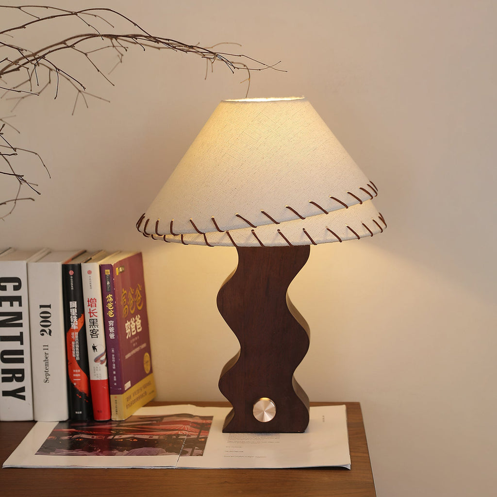 Clérachic table lamp glowing warm against a cream wall, with books and a magazine on the wooden shelf below it.