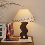 Clérachic table lamp glowing warm against a cream wall, with books and a magazine on the wooden shelf below it.