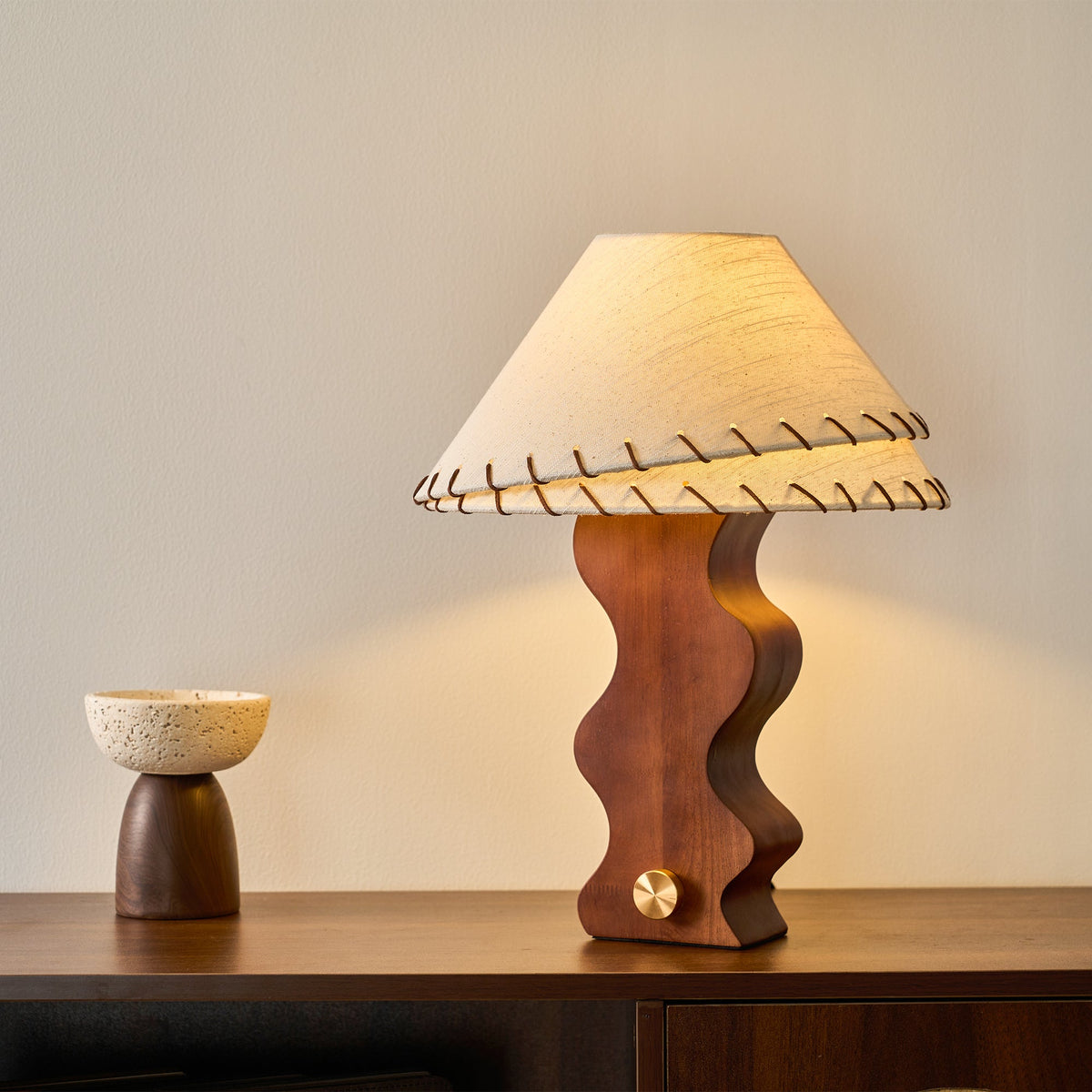 Lit Clérachic table lamp on a dark sideboard, its warm light falling on a small speckled bowl at the left.