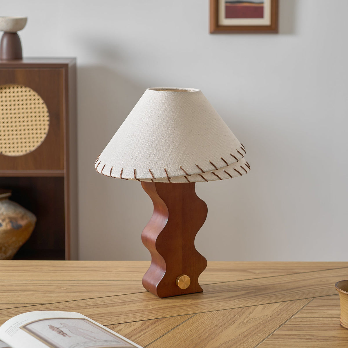 Unlit white linen shade on a Clérachic walnut lamp, set on an oak desk before a rattan-front cabinet and a vase.
