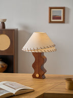 Portrait view of a Clérachic walnut table lamp on an oak desk, with a framed print on the grey wall behind.