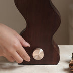 A hand turning the ribbed brass dial on the walnut base of this Clérachic table lamp, standing on pale stone.