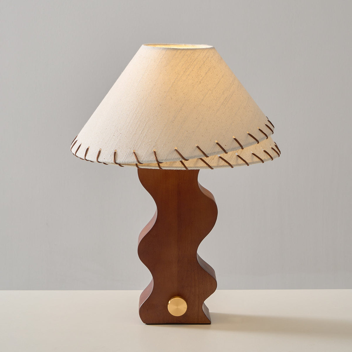 Clérachic table lamp lit from within, the linen shade glowing pale gold above the wavy walnut base and brass dial.
