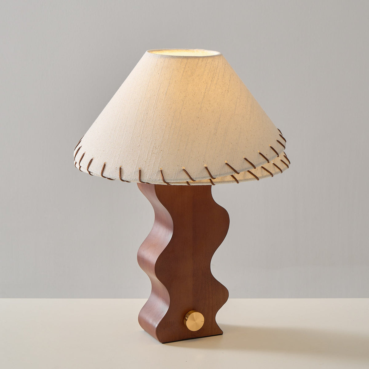 Clérachic walnut table lamp switched on, the glow lighting the underside of its stitched linen shade, against grey.