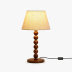 Walnut-toned stacked bead table lamp with a beige fabric shade from Clérachic, shown lit with its inline cord switch on white.