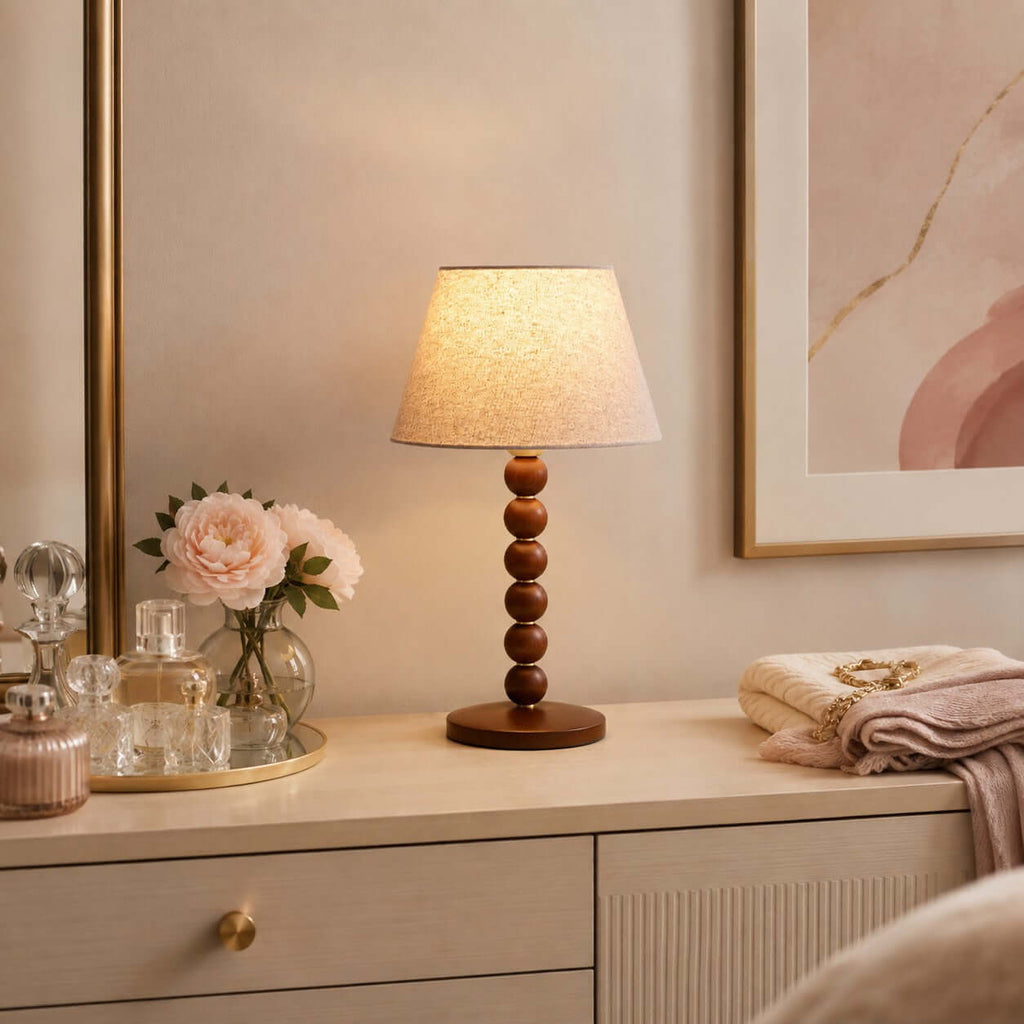 Lit Clérachic bead-column lamp on a cream dresser with pink roses, perfume bottles and a folded blanket beside it.